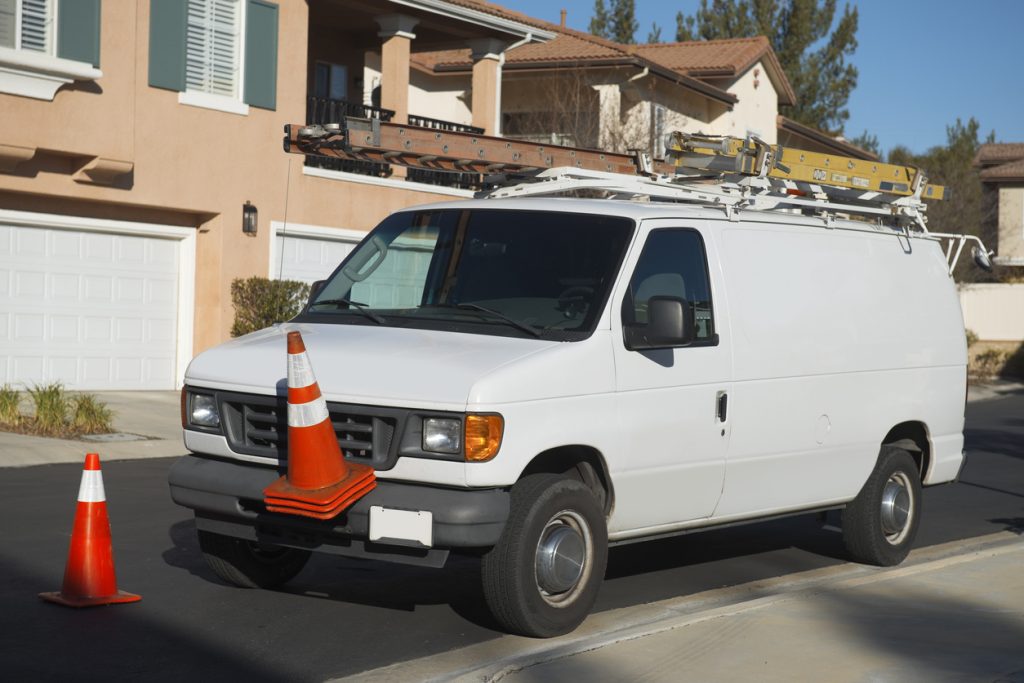 A white work van with ladders on its roof is parked on a suburban street. Orange traffic cones are placed in front of the van, with one cone resting on the front bumper. Houses and garages are visible in the background.
