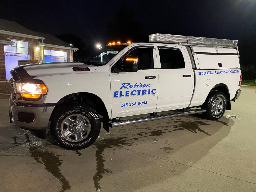 A white Robison Electric truck parked at night under bright lights. The truck displays company details and phone number, with a metal utility box on the bed and reflections on the wet pavement.