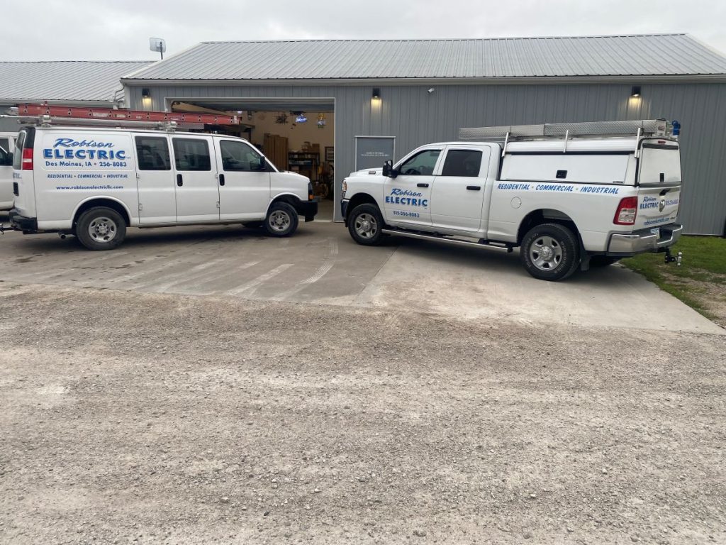 Two white Rohner Electric service vehicles are parked outside a gray metal building with an open garage door. The vehicles display the company’s logo, contact information, and a ladder is mounted on one van’s roof.