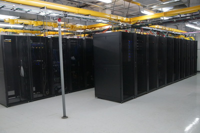 Rows of black server racks in a brightly lit data center, with yellow cable trays running overhead and white tile flooring.