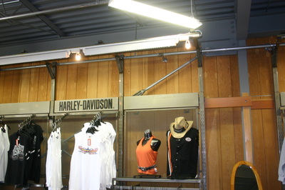 A retail display features Harley-Davidson apparel, including white T-shirts, an orange tank top on a mannequin, a black shirt, and a tan cowboy hat against a wooden wall. A Harley-Davidson sign is above the clothing.