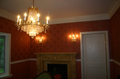 A warmly lit room with red patterned wallpaper, an ornate gold fireplace, a crystal chandelier, and wall sconces. A window is on the left and a white door is on the right.