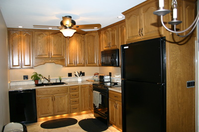 A kitchen with wooden cabinets, black appliances including a refrigerator, dishwasher, stove, and microwave, a ceiling fan with a light, and under-cabinet lighting. There are round black rugs on the tiled floor.
