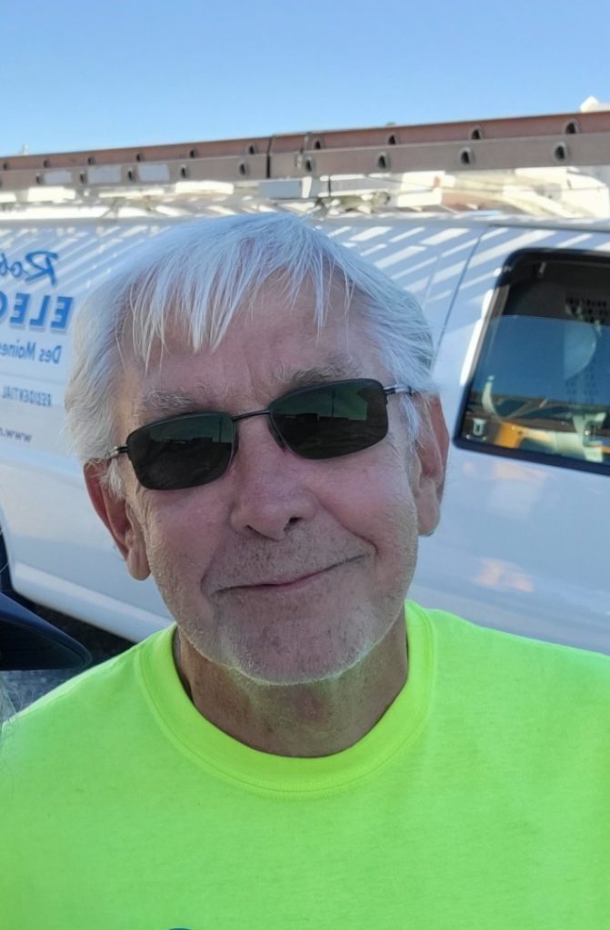 An older man with white hair and sunglasses smiles outdoors, wearing a neon yellow shirt. Behind him, there is a white utility van with a ladder on top and blue lettering on the side. The sky is clear and blue.
