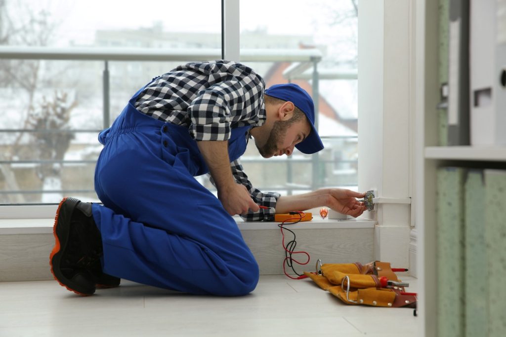 A man in blue overalls and a plaid shirt kneels on the floor, using a screwdriver to work on an electrical outlet. Tools and a tool belt are placed nearby. A large window shows an outdoor scene in the background.
