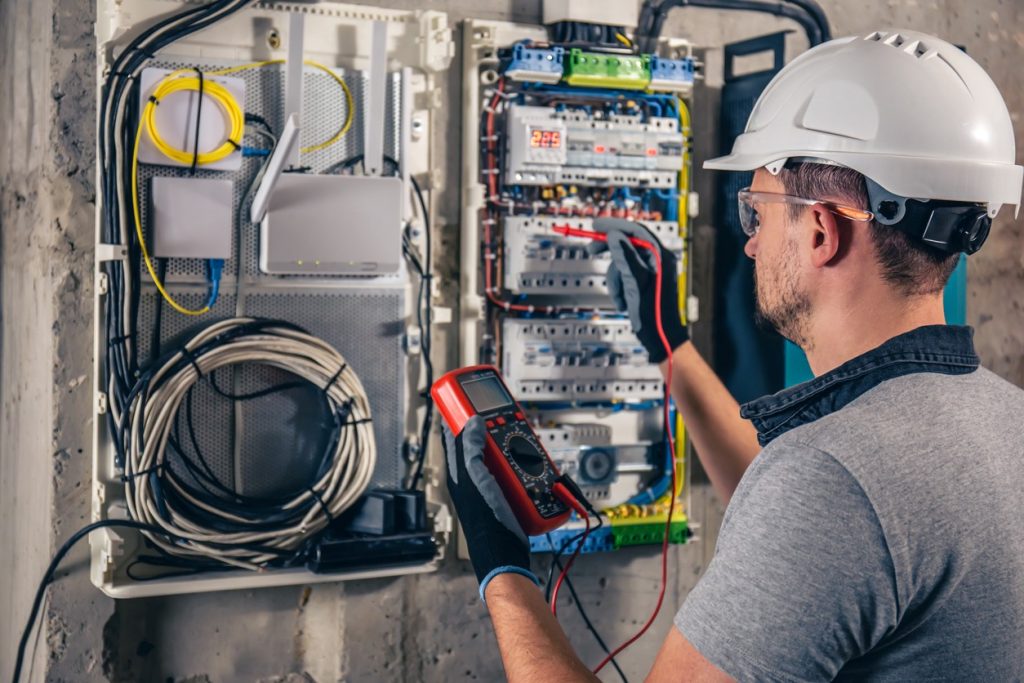 An electrician wearing a white hard hat and safety glasses tests an electrical panel with a multimeter. Network cables and a router are visible inside the open cabinet on the wall.
