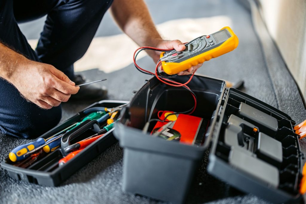 A person kneels on the floor, holding a yellow digital multimeter above an open toolbox filled with various tools such as screwdrivers, pliers, and wires.