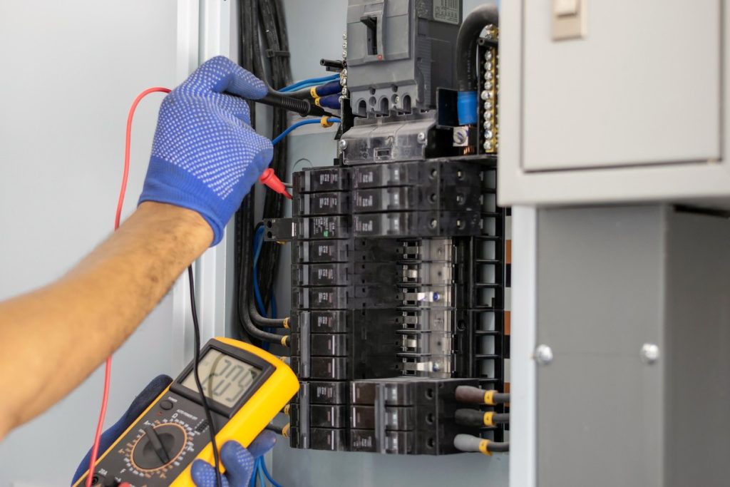 A person wearing blue gloves uses a multimeter to test wires inside an open electrical panel filled with circuit breakers.