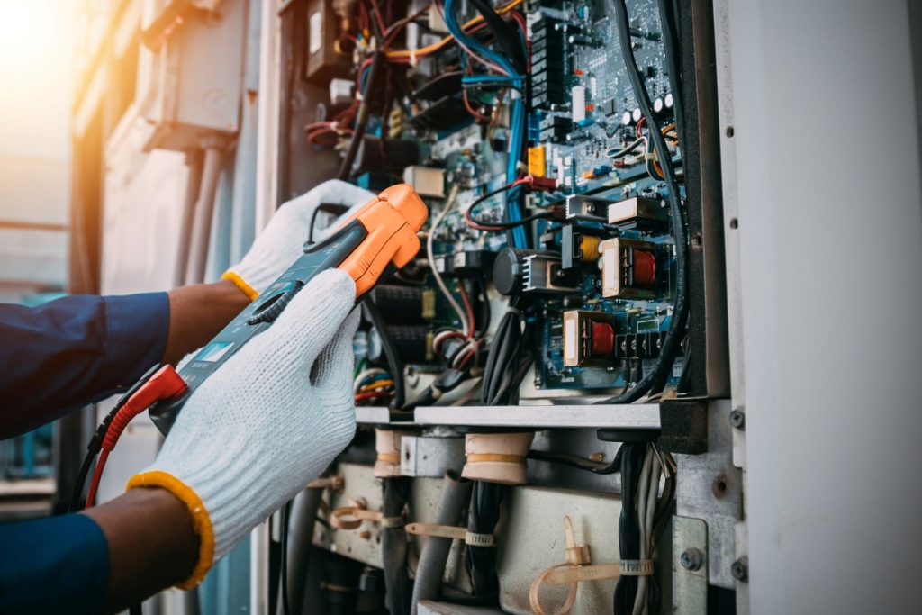 A person wearing gloves uses an electrical testing device on exposed wiring and circuits inside an open industrial control panel.