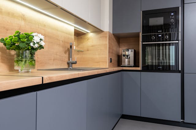 Modern kitchen with matte gray cabinets, wooden countertops and backsplash, built-in black oven and microwave, a stainless steel sink, a coffee machine, and a vase of green leaves with white flowers on the counter.