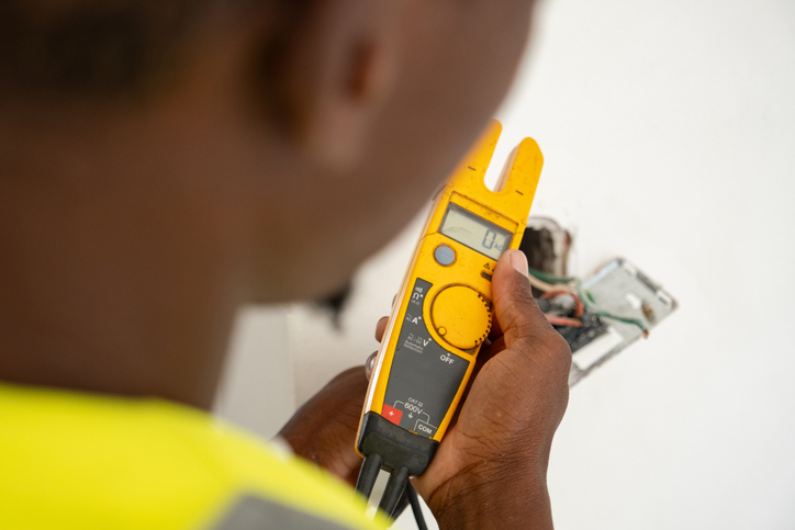 A person holds a yellow digital multimeter, testing electrical wires protruding from a wall outlet. The focus is on the tool and wires, with the person’s hand and part of their face visible in the foreground.