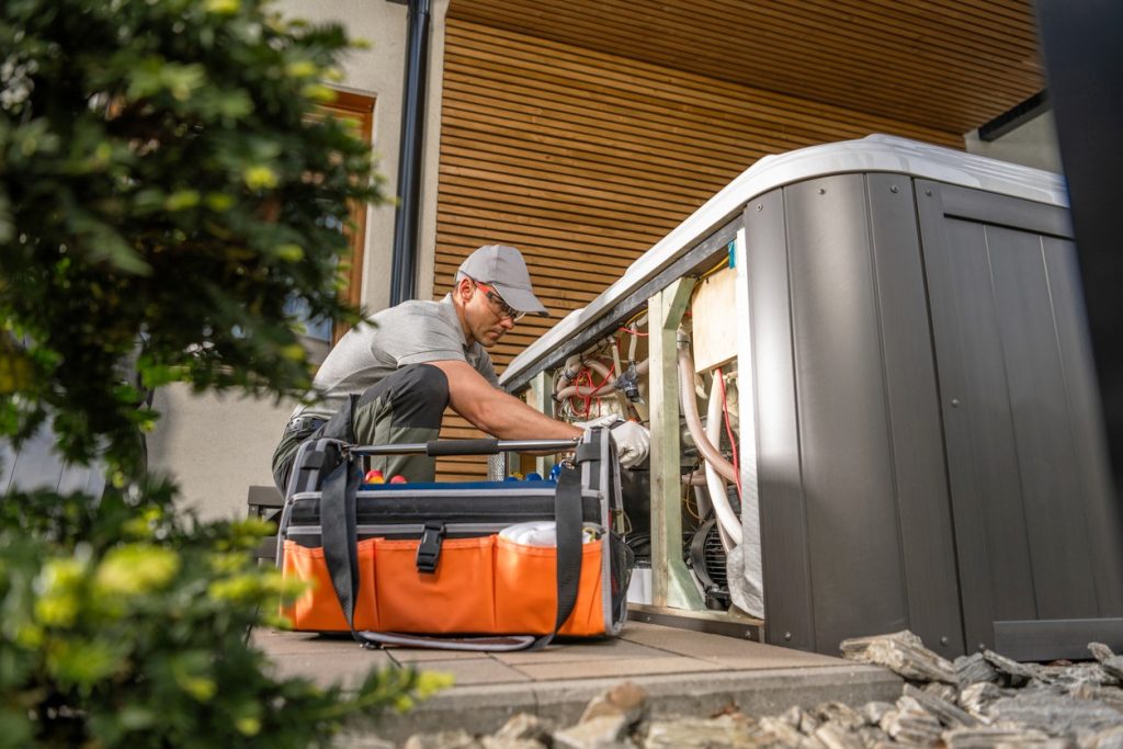 A technician wearing a gray cap and shirt is kneeling beside an open outdoor hot tub, working on its internal components. An orange tool bag sits on the ground nearby, with a shrub partially visible in the foreground.