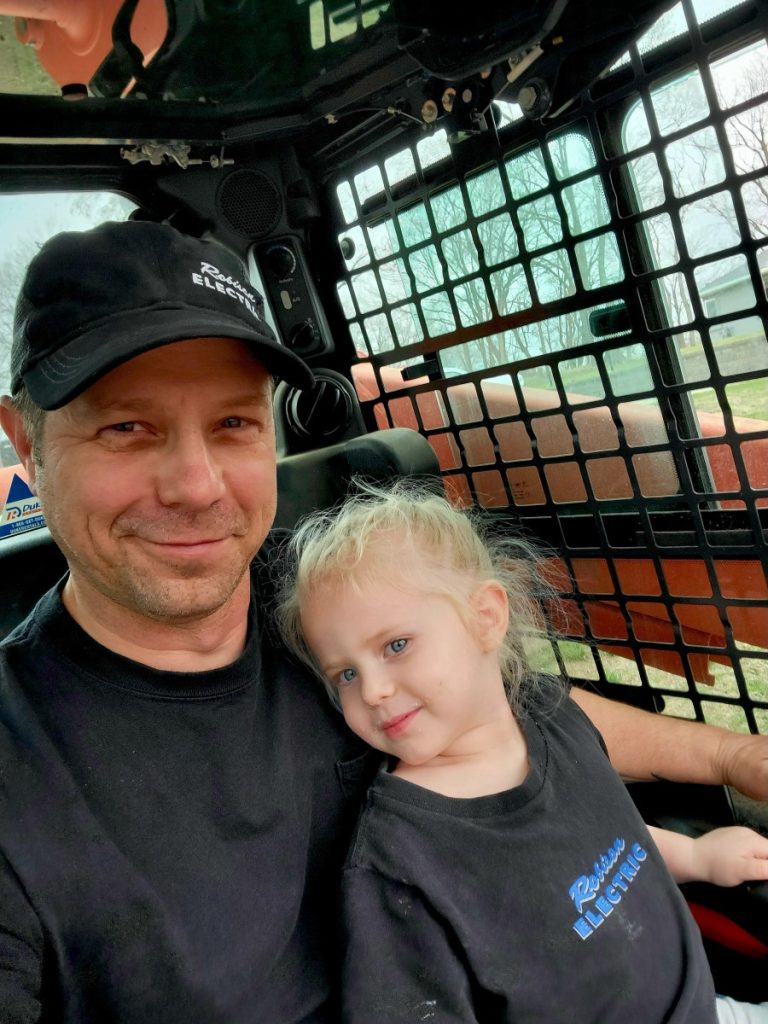 A man and a young girl, both wearing black shirts, sit together inside a vehicle with a metal safety grid. The man is smiling at the camera while the girl looks slightly to the side. Trees are visible outside the window.