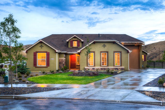 Single-story house with tan exterior, red shutters, and a brown roof, surrounded by a manicured lawn and wet pavement, under a partly cloudy sky at dusk. Warm lights shine from the windows.
