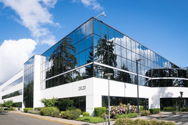 Modern office building with mirrored glass windows reflecting trees and sky, white exterior walls, and the number 15120 displayed on the front. Landscaping with bushes and a clear blue sky overhead.