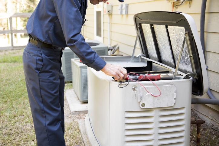 A technician in a blue uniform inspects or repairs an outdoor generator next to a house, using tools and testing equipment with the generator lid open.