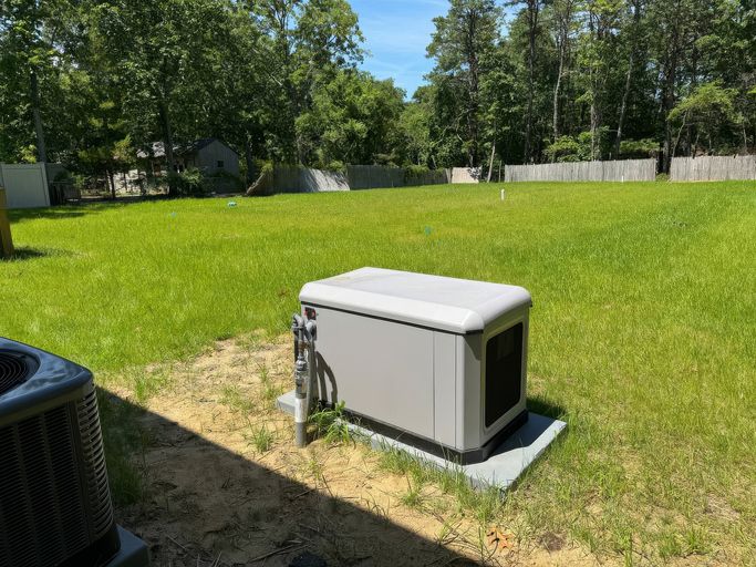 A standby generator sits on a concrete pad in a sunny backyard with green grass and trees in the background. A fenced area and some neighboring houses are visible in the distance.