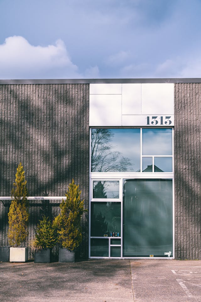 A modern gray building with the address number 1515 above a large windowed entrance, two potted trees on either side of the doorway, and a clear sky with clouds in the background.