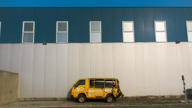 A small, damaged yellow van is parked against a large, plain white and blue industrial building with multiple rectangular windows. The van appears abandoned and has a dented rear.