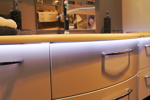 Close-up of modern bathroom cabinets with glossy white curved drawers, chrome handles, and soft LED lighting underneath the countertop. Reflections and decor items are visible in the background.