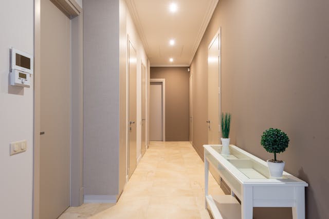A well-lit hallway with beige walls and floors, several closed doors, and a white console table holding two small potted plants. Ceiling spotlights provide illumination.