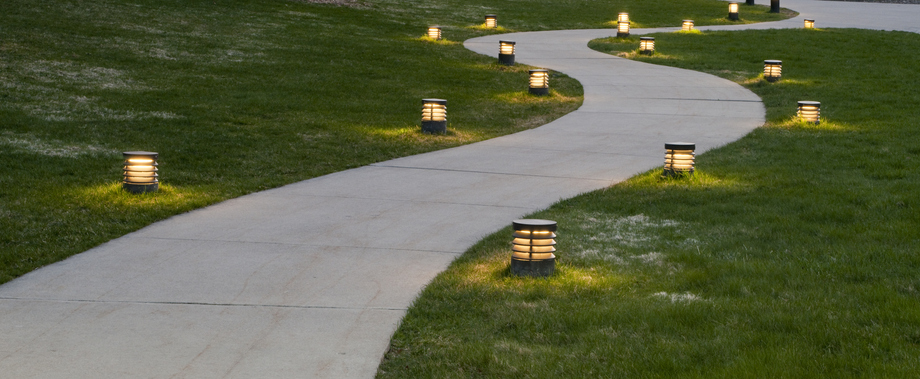 A winding concrete pathway through grass, lit by evenly spaced, round outdoor lights along both sides, glowing warmly in the evening.