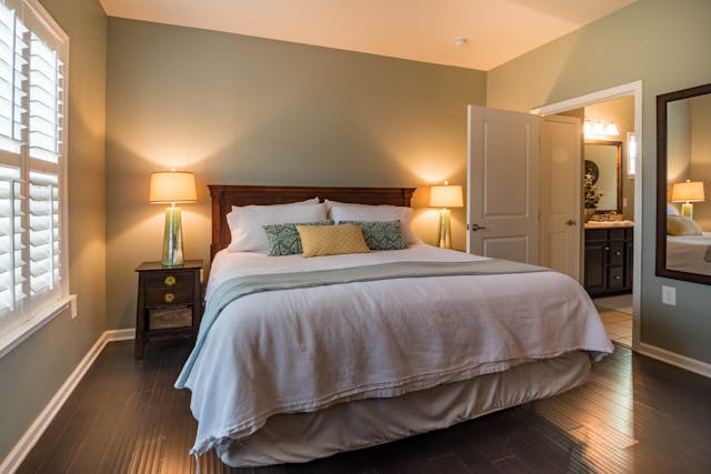 A cozy bedroom with a large bed, greenish walls, two bedside tables with lamps, a mirror on the right wall, and an open door leading to a bathroom. White shutters cover the window on the left.